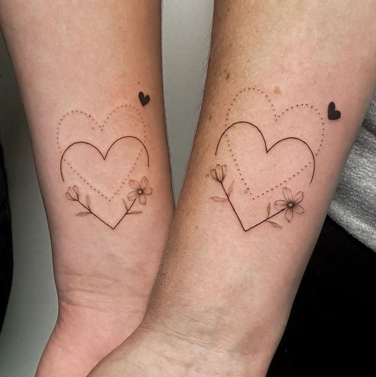 Two matching fine line heart tattoos with flowers and dotted hearts on the forearm