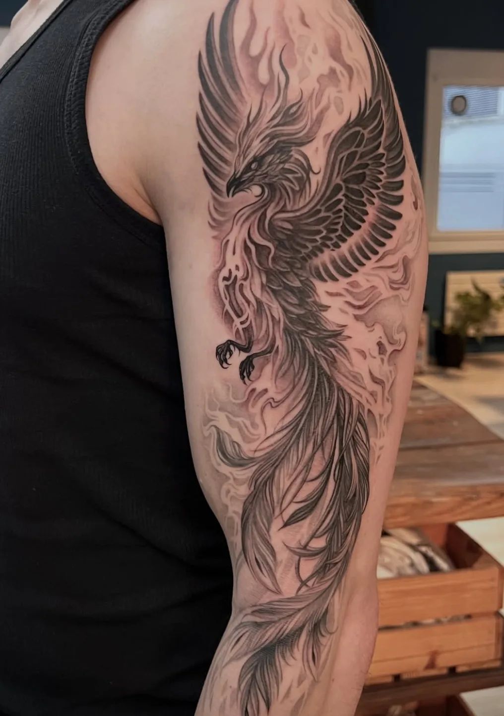 Phoenix tattoo in black and grey style on upper arm with flames and smoke