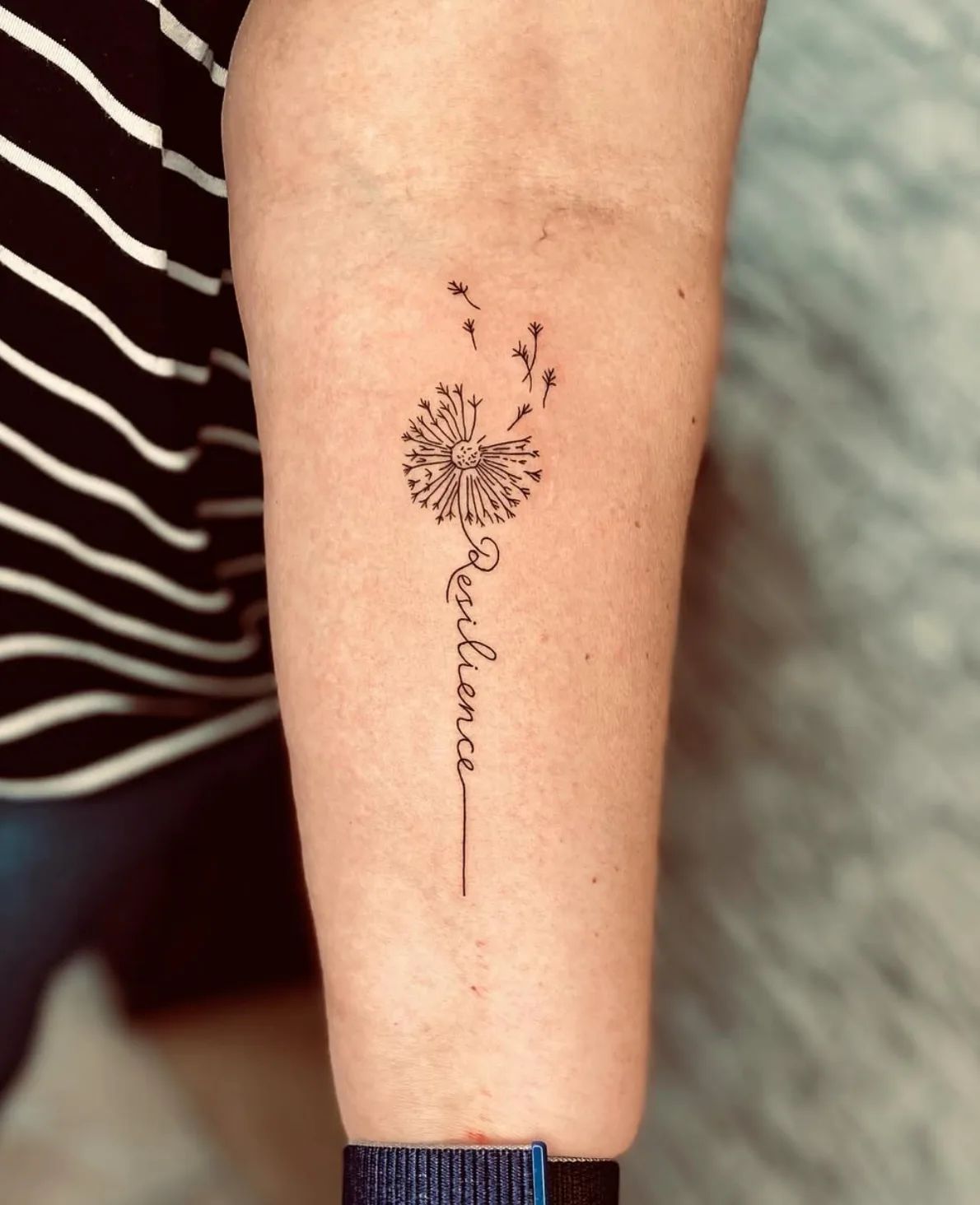 Fine line dandelion tattoo with "Resilience" lettering on upper arm