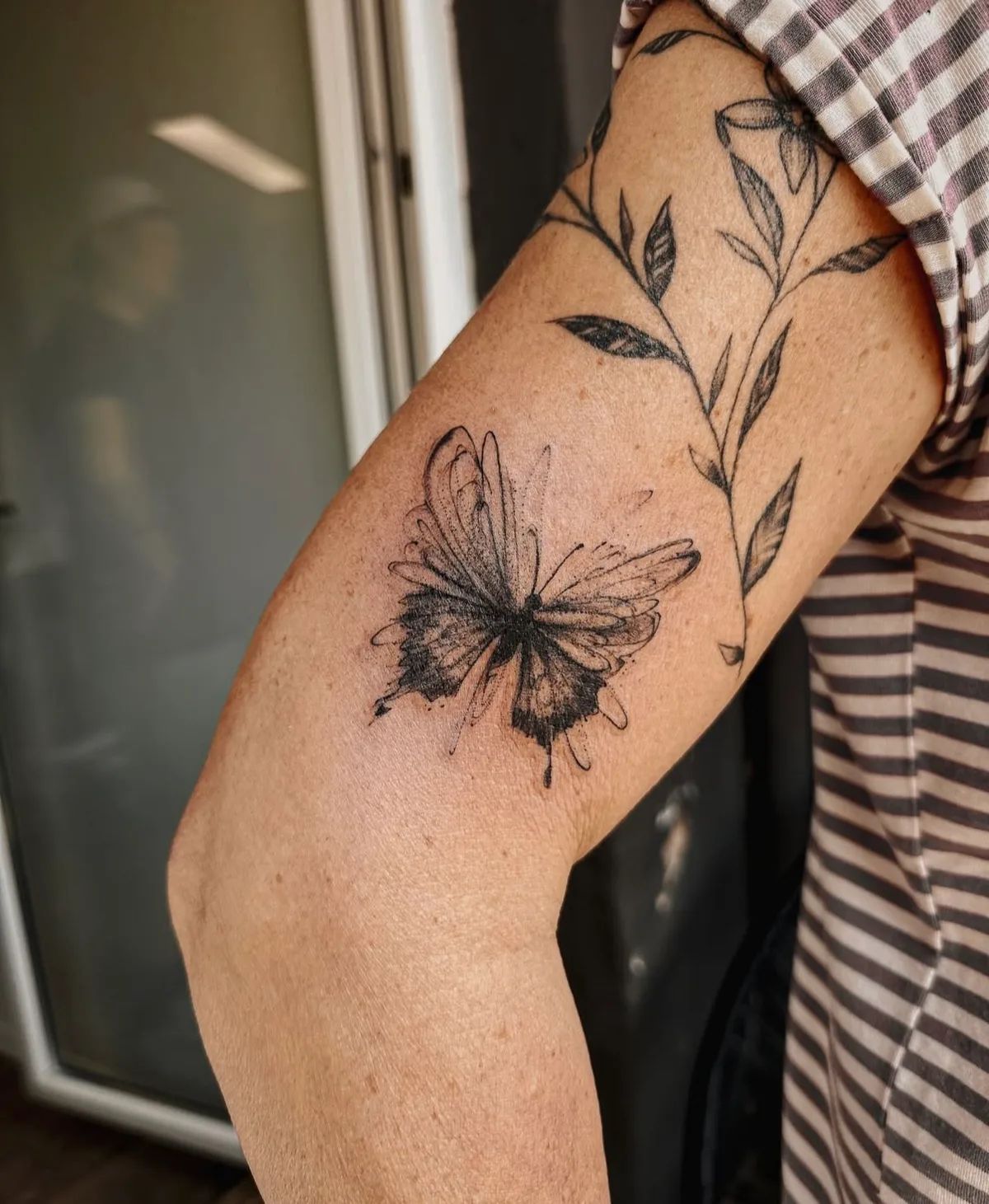 Schmetterling Tattoo im Fineline-Stil mit Blättern am Oberarm
