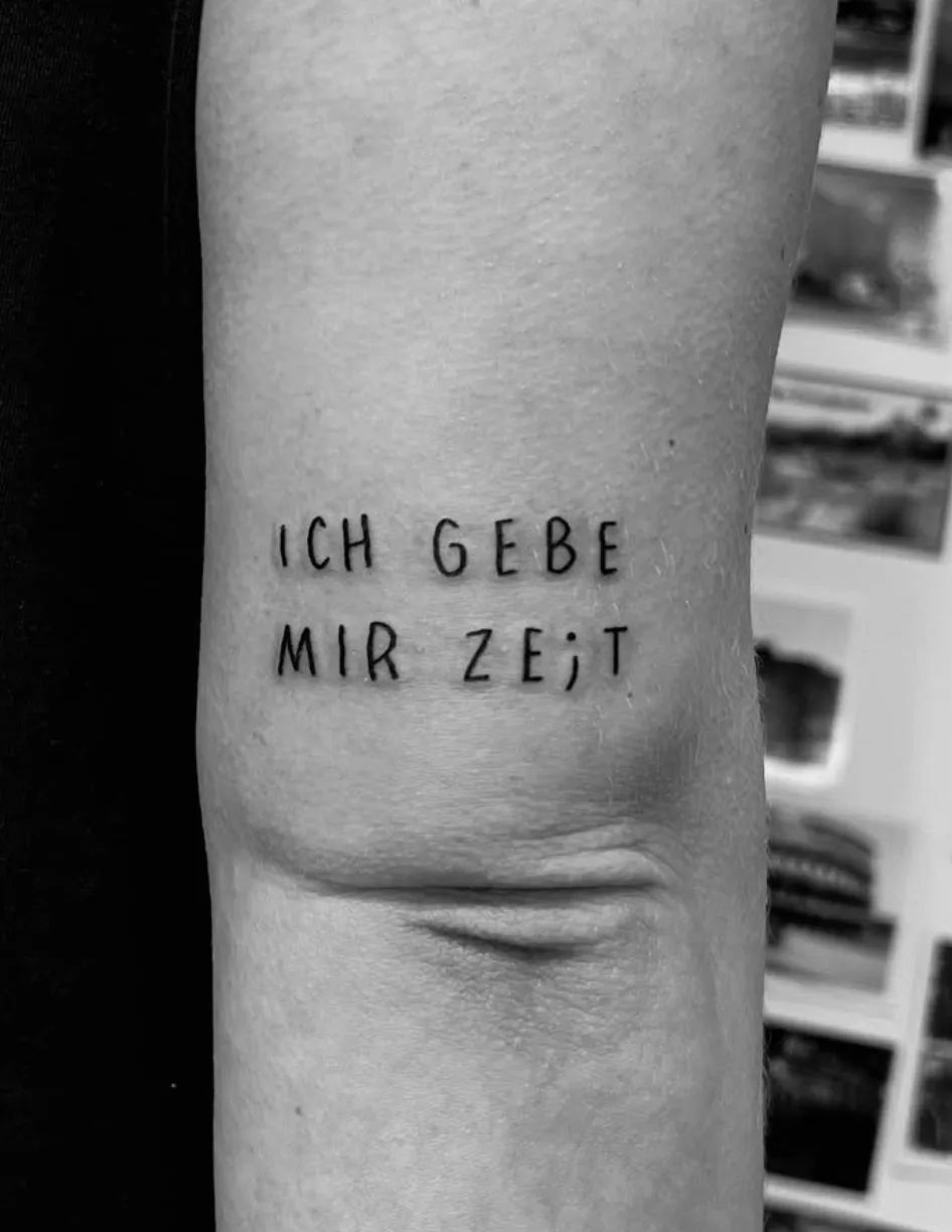 Lettering tattoo "ICH GEBE MIR ZE;T" in fineline script on upper arm
