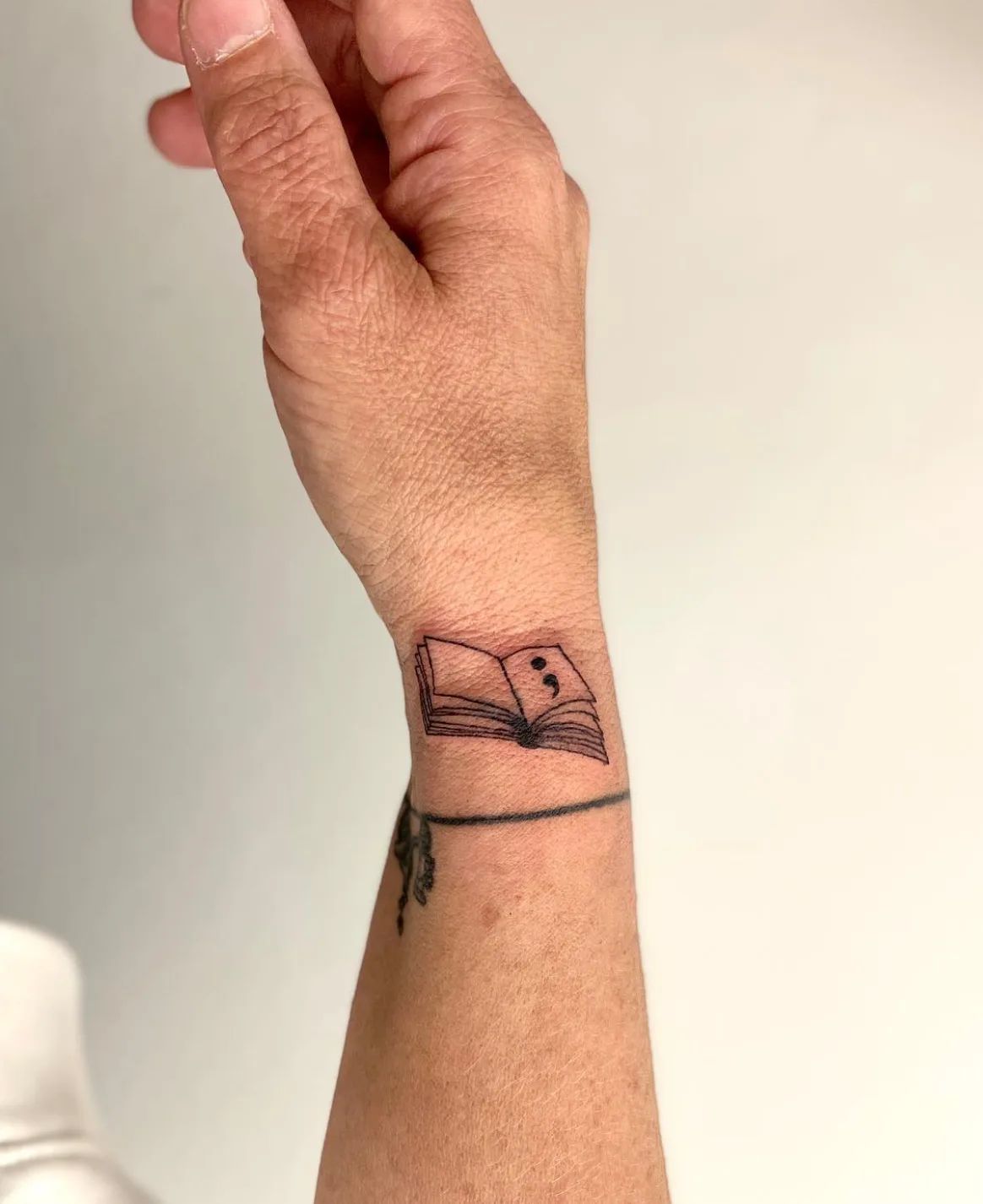 Semicolon tattoo as an open book in fine line style on the wrist