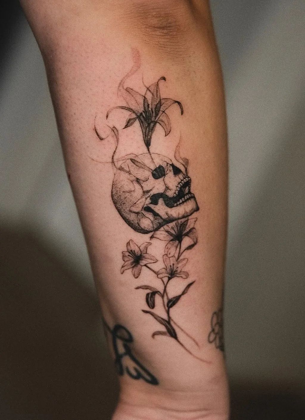 Skull tattoo with lilies and smoke in fine line style on forearm