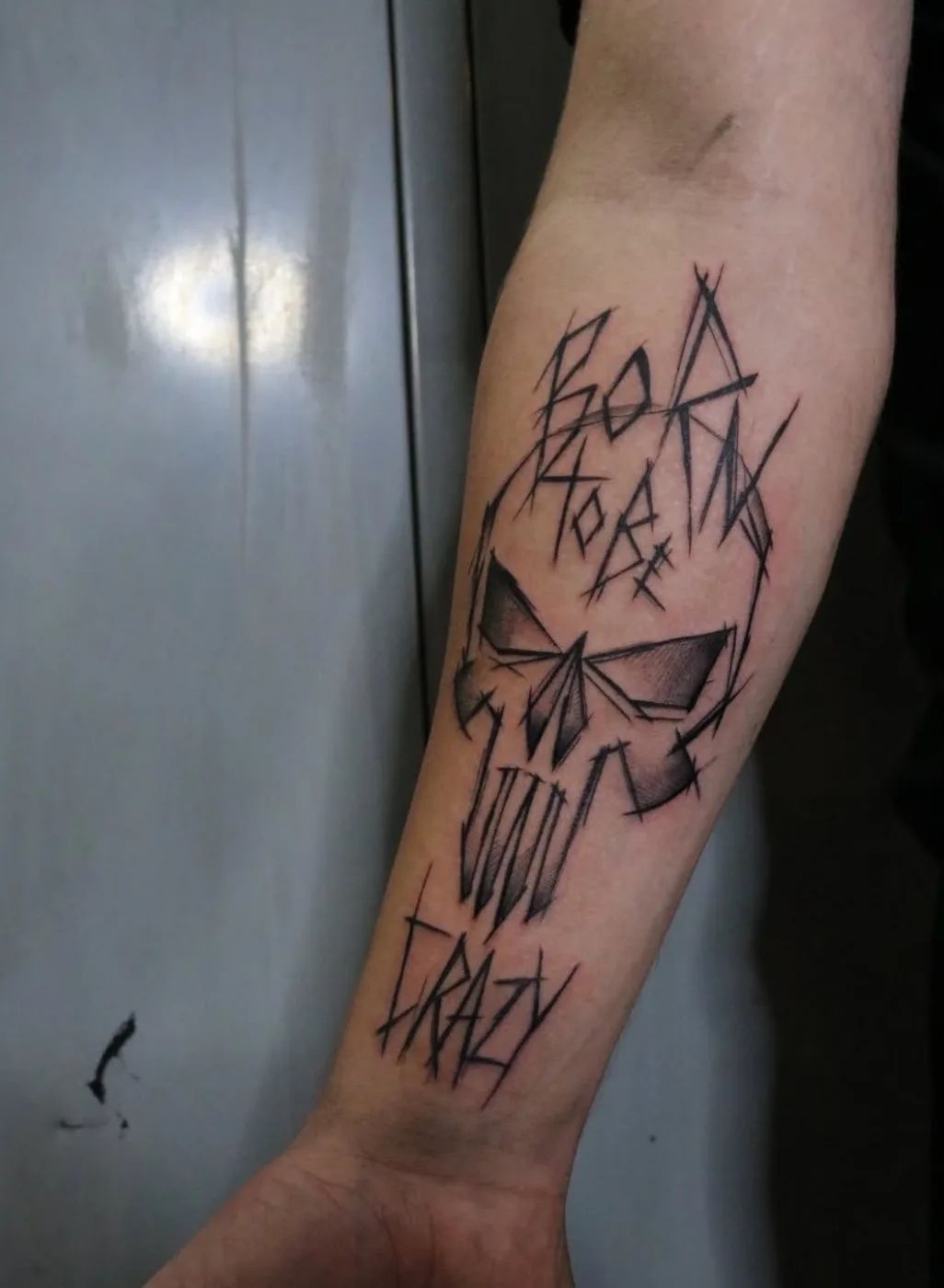 Skull tattoo in trash polka style with "Born to be crazy" lettering on forearm